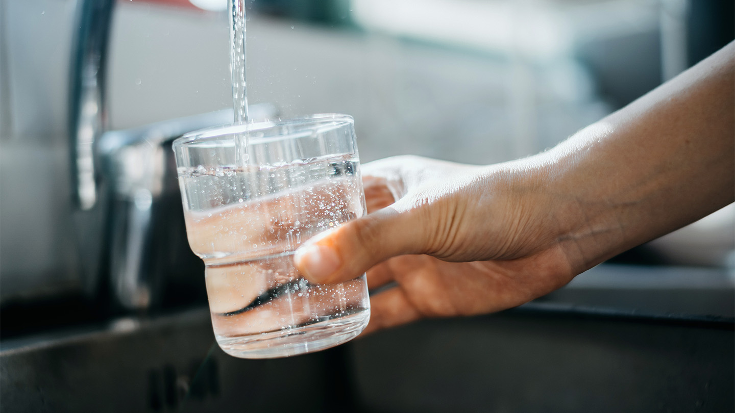 A stock image of a person holding a glass under a faucet. The cup is being filled with drinking water.